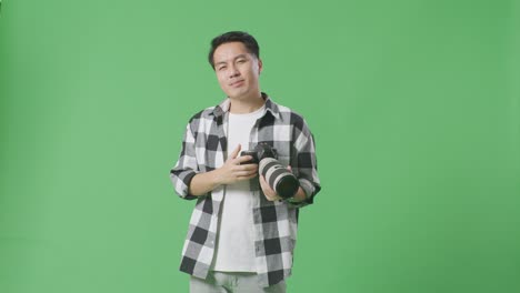 asian photographer using a camera taking pictures and making shh gesture while standing on green screen background in the studio