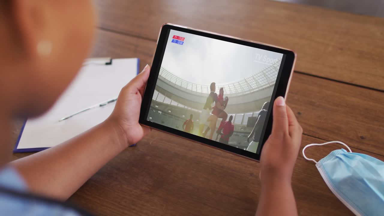 태블릿에서 럭비 경기를 시청하는 얼굴 마스크와 함께 테이블에 앉아있는 여성의 복합