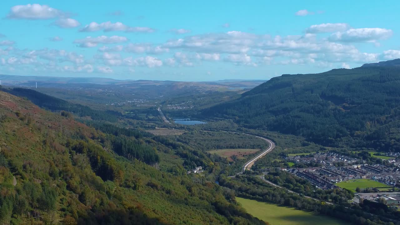 Retreating Aerial Over Welsh Landscape with Rugged Natural Forest Hills and Winding Heads of the Valleys Road with Rural Houses and Blue Sky