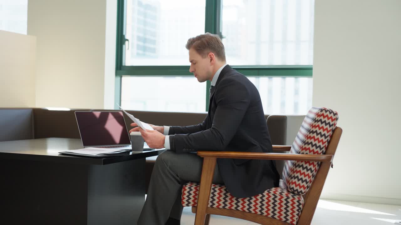 A professional in a dark suit sits in a mid-century modern chair with chevron upholstery, working on a laptop and reviewing documents while taking a coffee break in office space with large windows