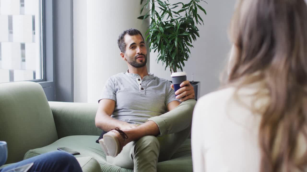 En forretningsmand af blandet herkomst sidder på en sofa, holder en kop kaffe og taler med kolleger.