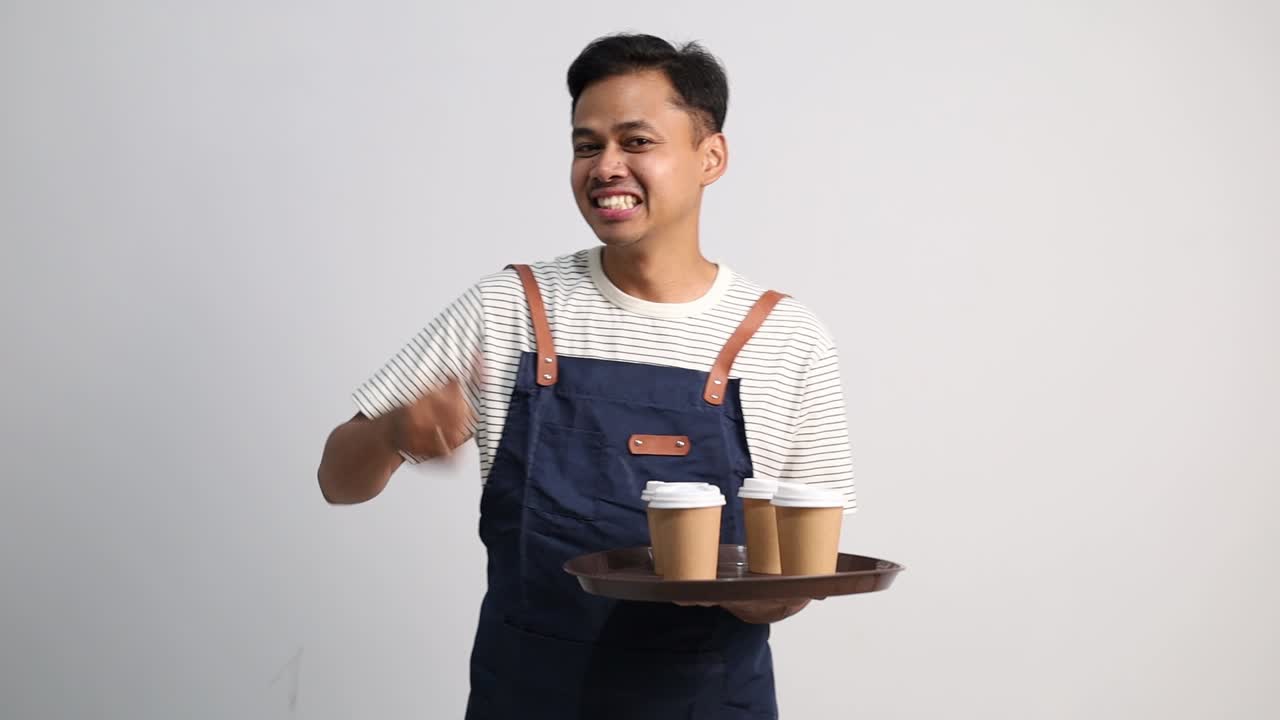 Seorang pria muda mengenakan celemek menunjuk ke nampan berisi gelas kertas, mempromosikan atau menyajikan produk minuman dan memberikan jempol.
