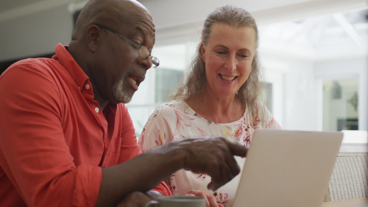 Happy senior diverse couple wearing shirts and using laptop in living room