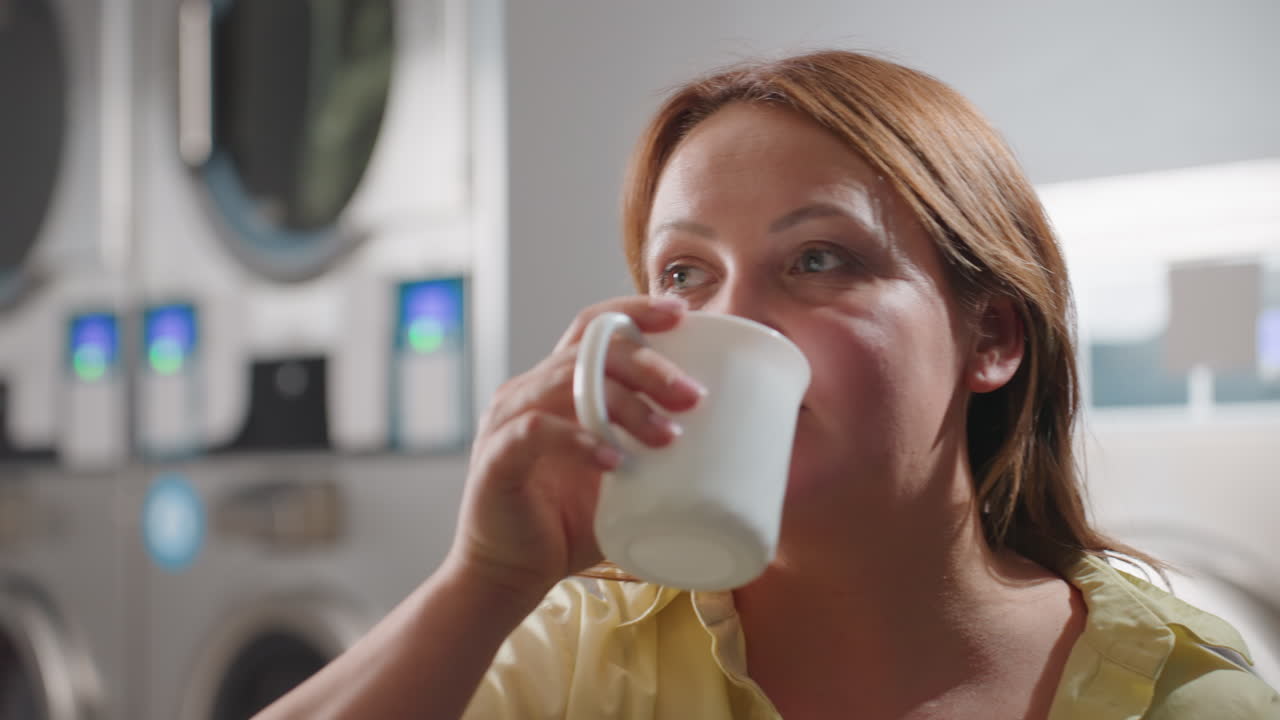 Close up of female worker seated inside laundromat drinking hot beverage from white mock cup, industrial washing machines glowing in background, break time relaxation, hygiene, modern lifestyle