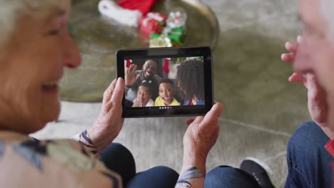pareja de ancianos caucásicos sonrientes usando una tableta para una videollamada de navidad con la familia en la pantalla