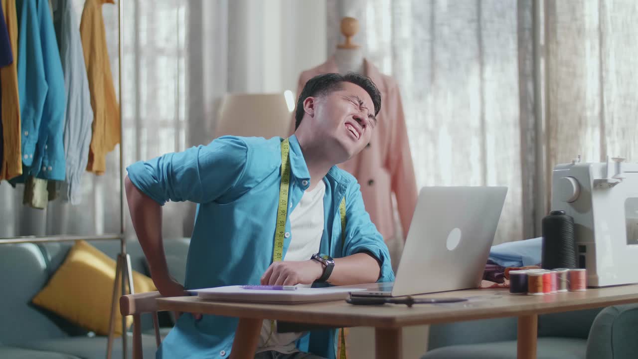 아시안 남성 디자이너가 노트북으로 작업하고 스튜디오에서 옷을 디자인하는 동안 허리 통증이 있습니다.