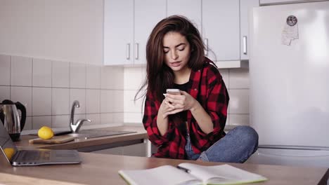 hermosa chica morena enviando mensajes de texto a alguien mientras estaba sentada en la cocina de su apartamento.