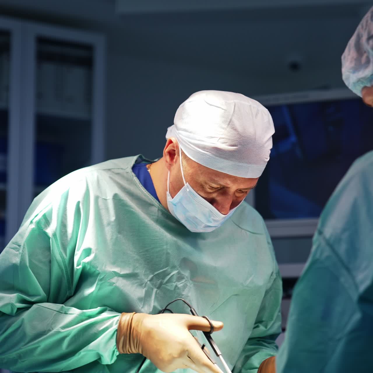 Experienced plastic surgeon using electric scissors instrument at surgery. Male doctor assisting the professional. Blurred backdrop