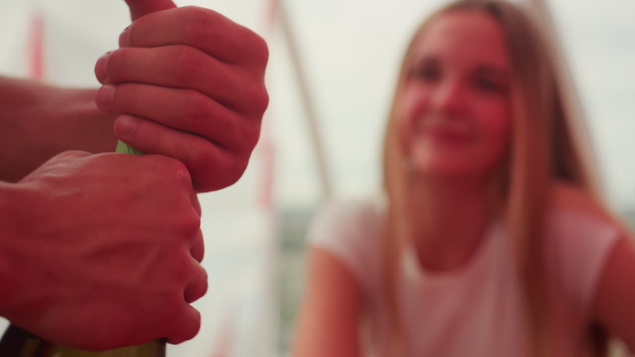 Giovane donna bianca che guarda lo champagne che spumeggia su una terrazza mentre il partner apre la bottiglia, mani che rimuovono il tappo in primo piano, risata sorpresa e calda luce del tramonto, abbigliamento estivo casual, momento gioioso e spontaneo