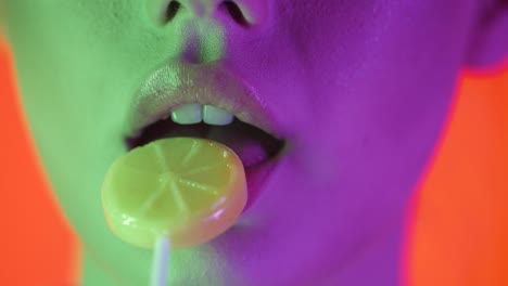 estrecha toma de cerca de los labios de una mujer joven y sexy mientras gira una dulce y deliciosa piruleta en forma de limón contra un fondo naranja con contraste verde púrpura en la cara en cámara lenta