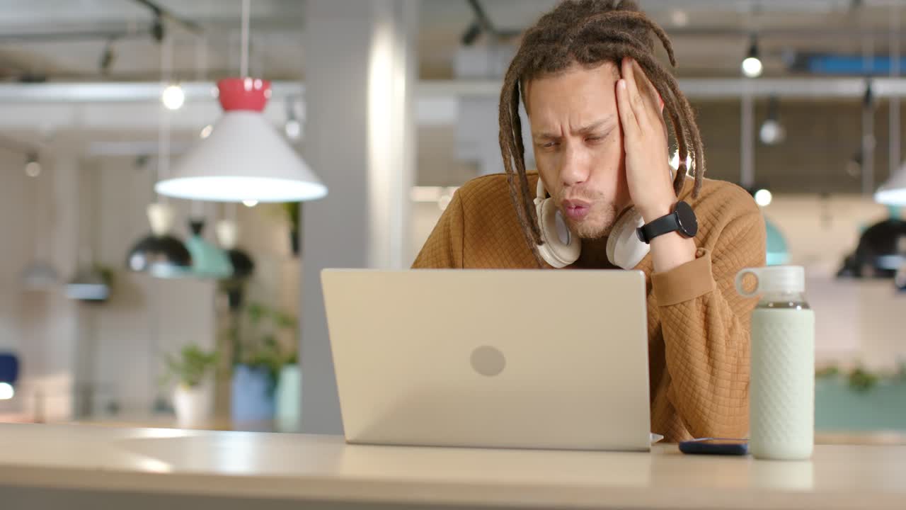 African American man leaning at coworking desk, studying laptop, drinking, rubbing cheek from pain