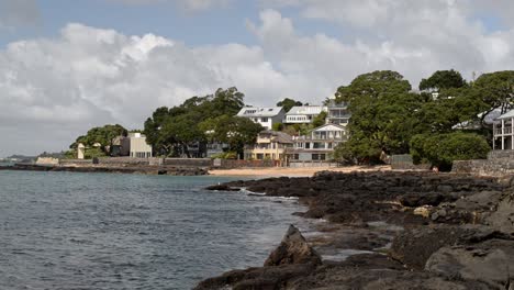 Several-beach-cottages-on-the-volcanic-north-shore-of-Auckland,-New-Zealand-on-a-sunny-day-with-big-cumulus-clouds-above