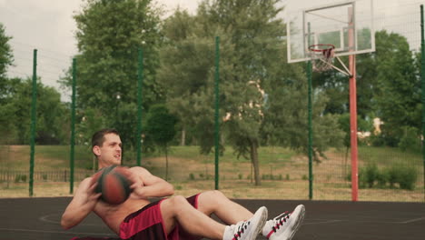 handsome shirtless sportsman doing abdominal exercises, holding a basketball ball and lying at ground in an outdoor basketball court