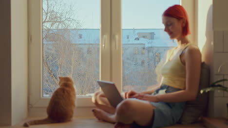 girl working on laptop on windowsill at home, her cat resting nearby