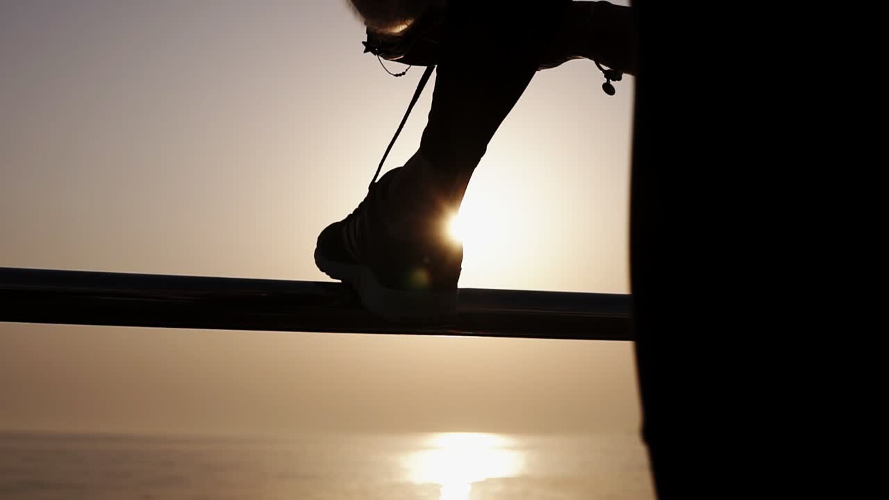 Extremely close up. Silhouette of a slender girl, ties up shoelaces on sneakers leaning on the crossbar. Morning sunrise on the background