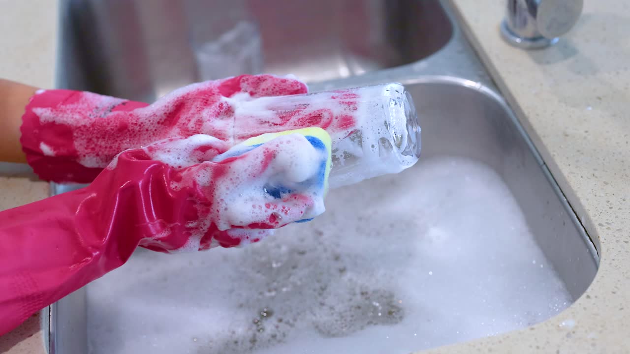 Hands in pink gloves wash a glass with a sponge in a kitchen sink, surrounded by soapy water