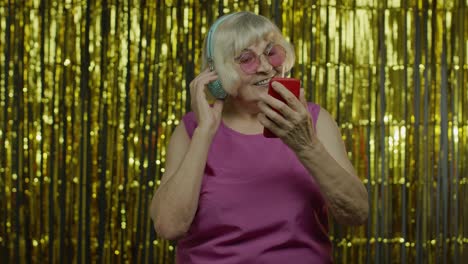 Stylish-happy-senior-old-woman-holding-cell-phone,-listening-to-music-in-headphones-and-dancing