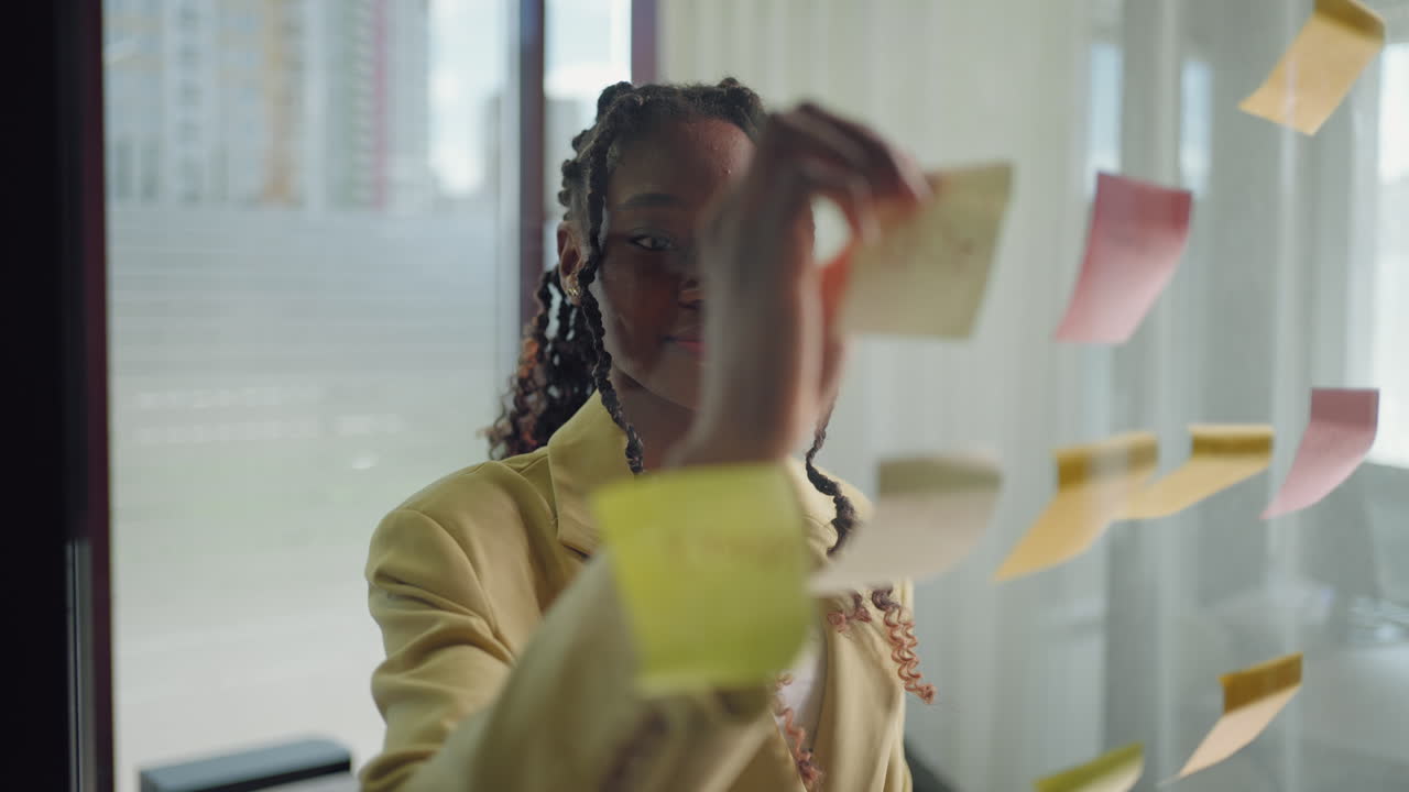 African american business woman using sticky notes brainstorming problem solving strategy on glass whiteboard leader woman showing solution for project deadline in office. Bringing her vision