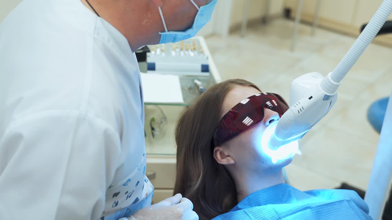 A woman at the dentist in red UV protective glasses is undergoing laser teeth whitening. A dentist in a blue mask is watching a patient