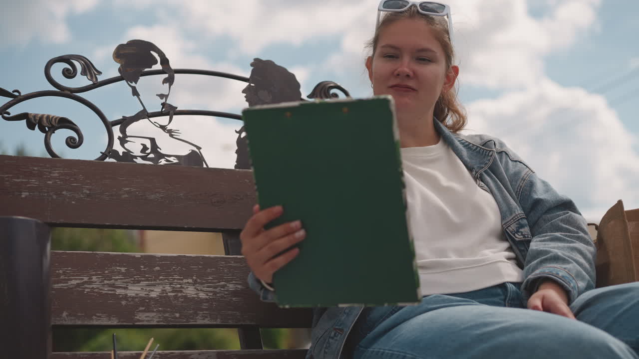Young white woman reading clipboard outdoors, sitting on weathered iron bench under blue sky, wearing denim jacket and sunglasses, flipping through green folder, relaxed expression, soft sunlight