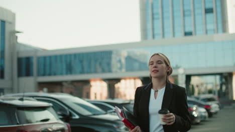 focused professional woman walking with folder drinking coffee among parked cars in urban setting with glass office building background on sunny day