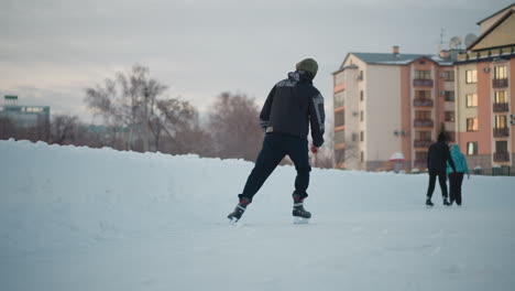 experienced boy skating skillfully on frozen lake wearing patterned jacket and hoodie near snow banks blurred background of novice skaters and apartment buildings under soft winter sky