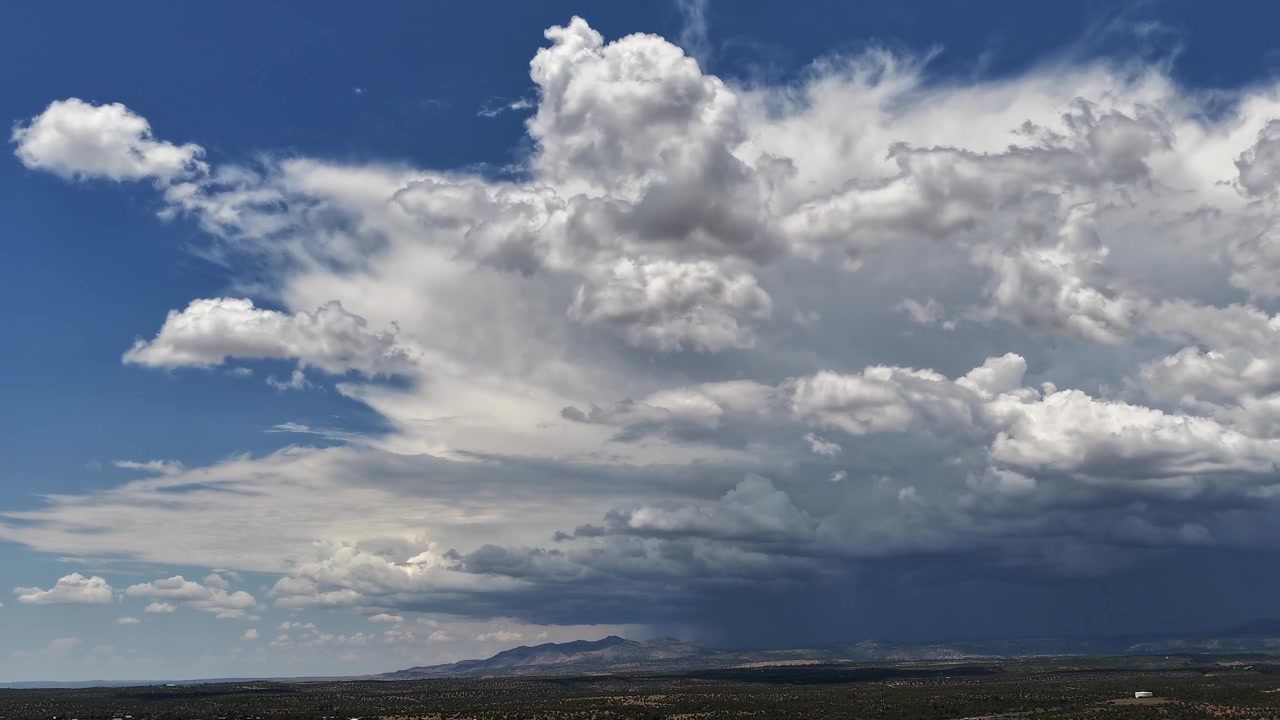 4K drone-timelapse, der fanger en massiv monsunstormdannelse. Flere lag af hurtigtbevægende, tunge cumulonimbus-skyer bygger sig dynamisk op over et stort åbent landskab mod en blå himmel.