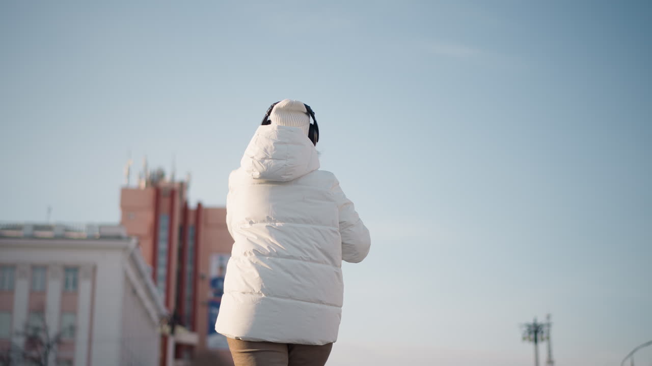 Cheerful dancer wearing white winter coat and beanie moves rhythmically in open urban space, back turned to camera, enjoying winter atmosphere under clear sky with city buildings in background