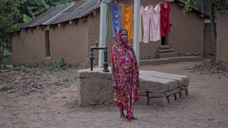 indian woman wearing a traditional sari stands near a water pump in a rural village, clothes hanging to dry in the background, showcasing the simplicity of village life