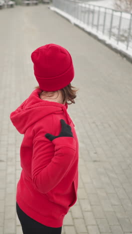 lady performing hand stretch by turning her arm outdoors in winter fitness routine, dressed in athletic wear, snow-covered ground, frosted trees, benches in serene park setting