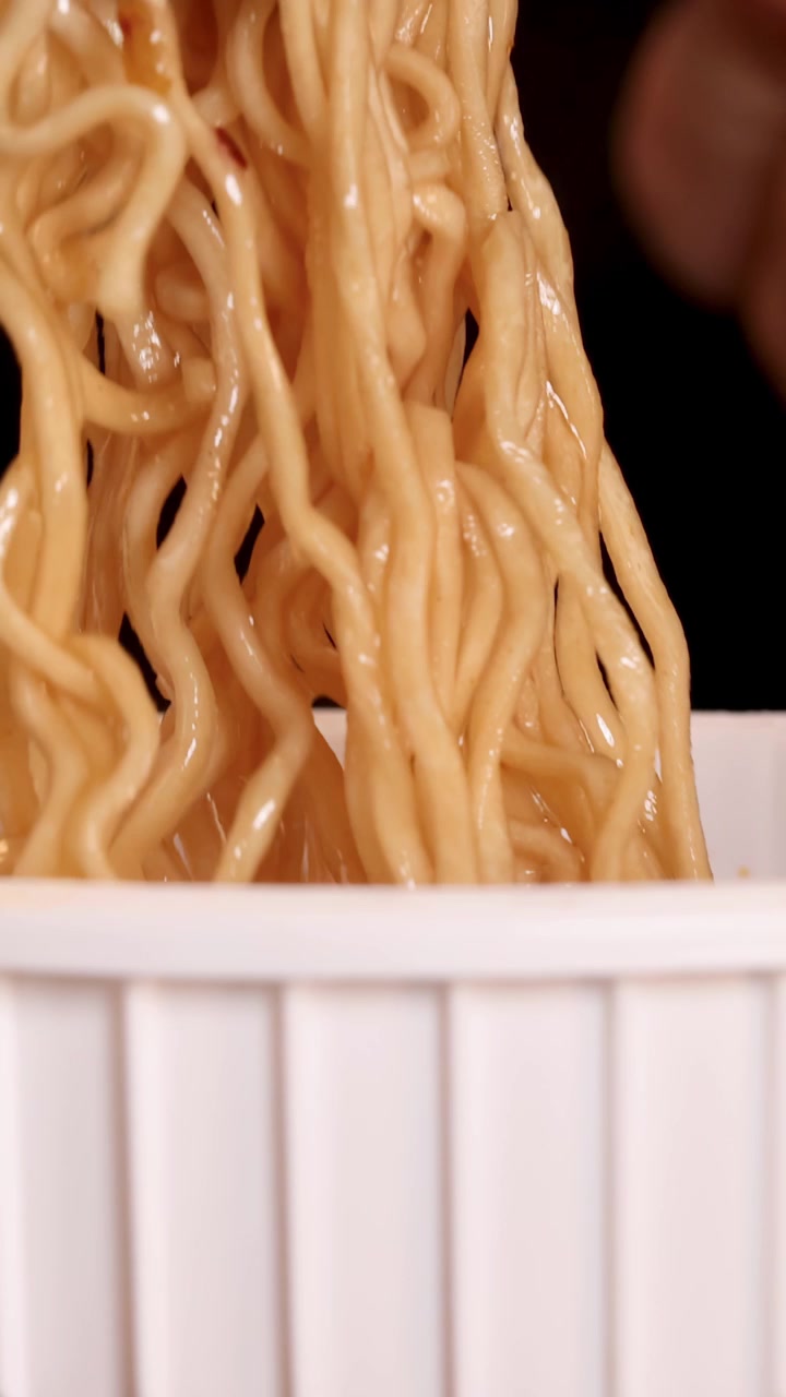 Una mano usa bacchette di legno per prendere noodles istantanei cotti da una tazza bianca a coste sotto un'illuminazione calda e focalizzata con uno sfondo scuro