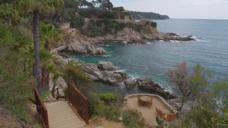 Cala-Banys-In-Lloret-De-Mar-Blick-Auf-Den-Felsstrand-Mittelmeer-Transparentes-Türkisblaues-Wasser-Spektakuläres-Restaurant-Direkt-Am-Meer-Mit-Chill-out-stühlen-Und-Direktem-Blick-Auf-Die-Klippen