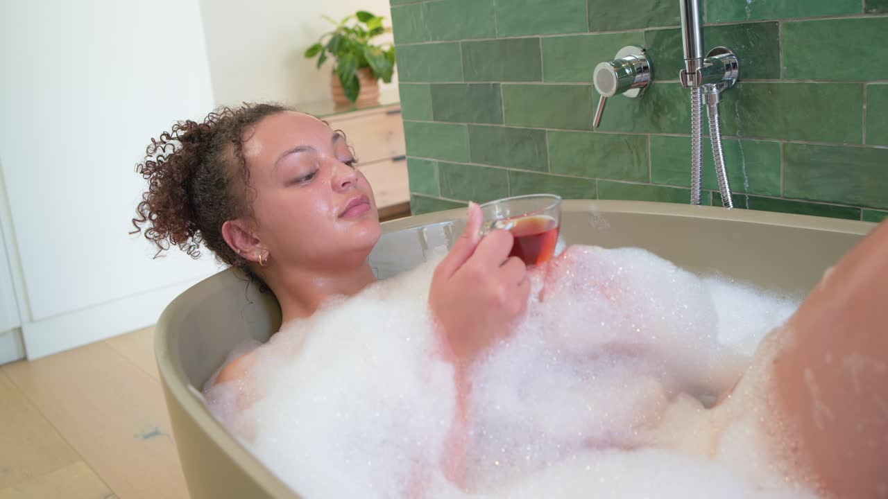 With camera moving, African American woman reclining in tub, sipping glass cup, relaxing
