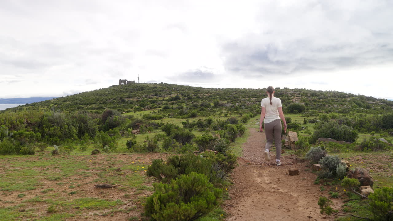 Veduta posteriore di una giovane donna che cammina su un sentiero sterrato attraverso l'Isla del Sol, Lago Titicaca, Bolivia, esplorando un paesaggio di collina verdeggiante sotto un cielo diurno nuvoloso.