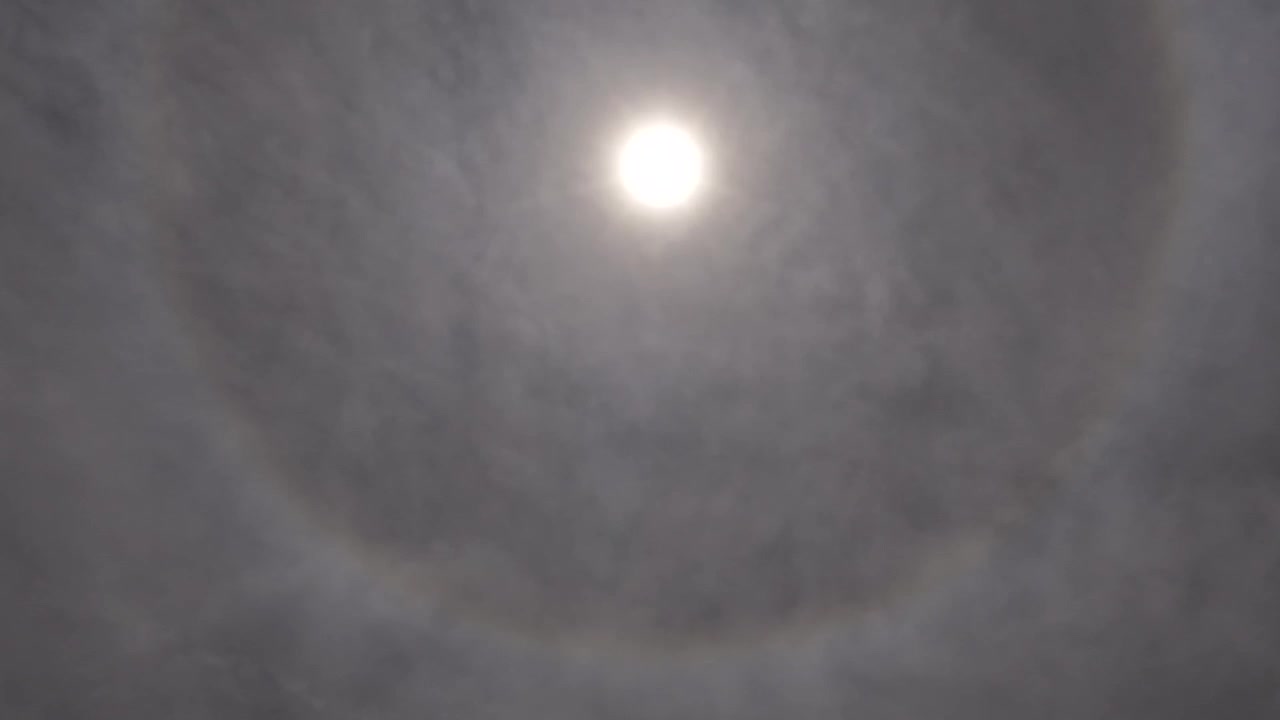 Un timelapse meteorologico diurno che mostra un prominente alone solare ottico. Uno strato di nubi cirri ghiacciate e in rapido movimento si sposta velocemente attraverso il sole splendente, evidenziando complesse dinamiche atmosferiche