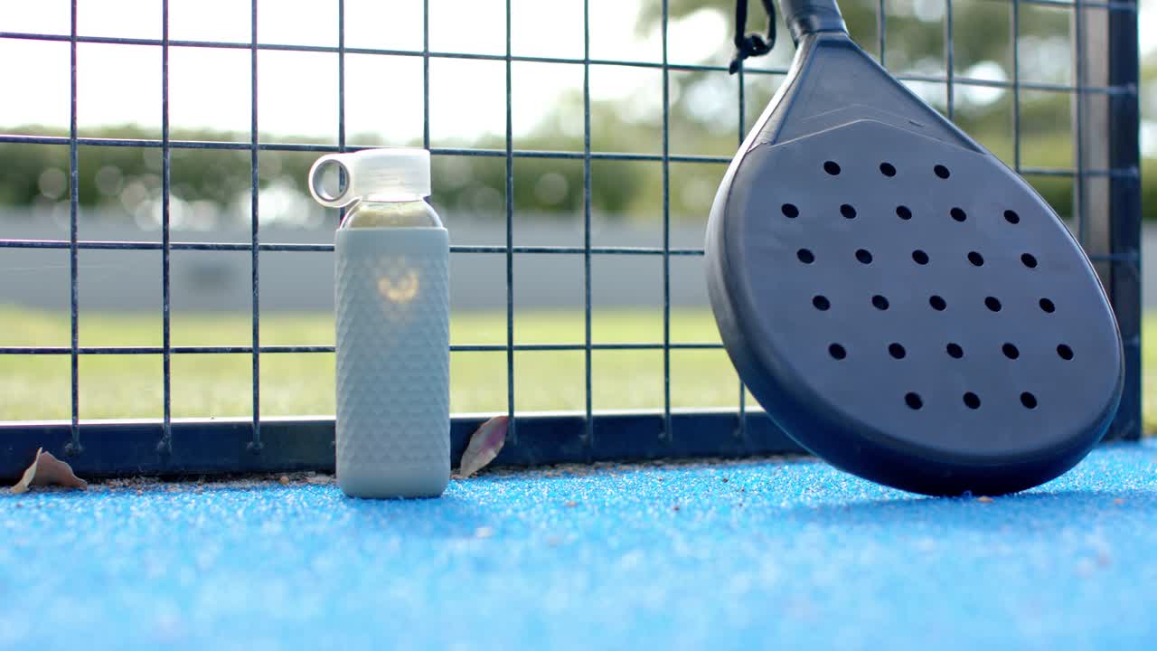 Neon tennis ball entering from right, bouncing, rolling past water bottle to paddle on blue court