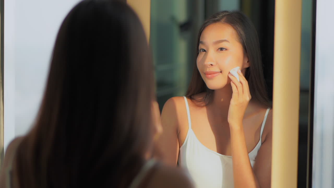 Young Asian woman cleaning the face with cotton pad near a mirror, handheld closeup