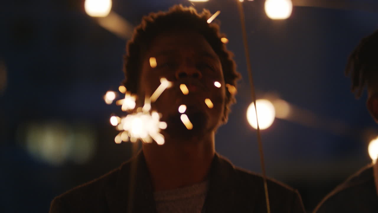 giovane uomo afroamericano che tiene le stelle filanti che ballano festeggiando il capodanno sul tetto di notte divertendosi con gli amici godendosi la celebrazione della festa