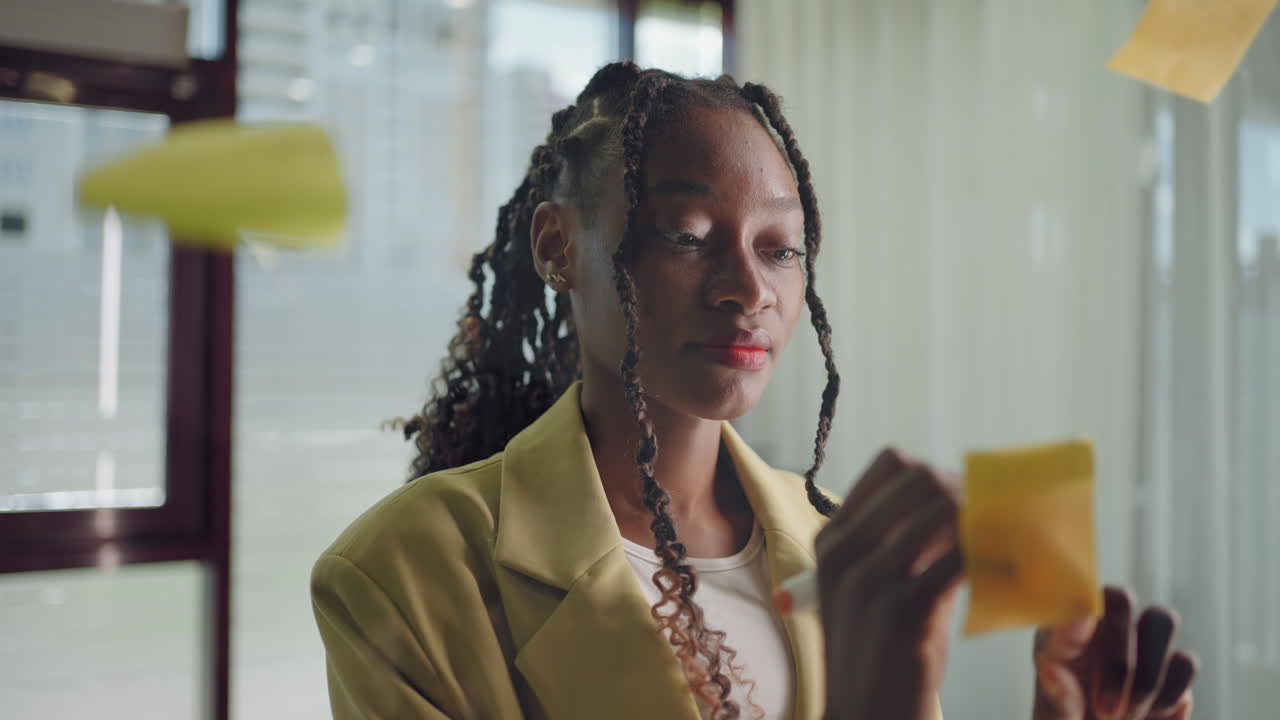 African american business woman using sticky notes brainstorming problem solving strategy on glass whiteboard leader woman showing solution for project deadline in office.