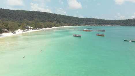 Beautiful-Turquoise-Blue-shallow-shore-extending-along-the-coast-with-fishing-boats-moored---Aerial-low-angle-panoramic-shot