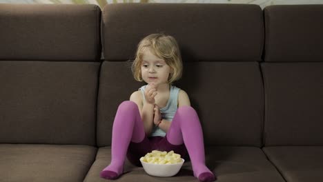 girl sitting on sofa and eating corn puffs. child smiling and taste puffcorns