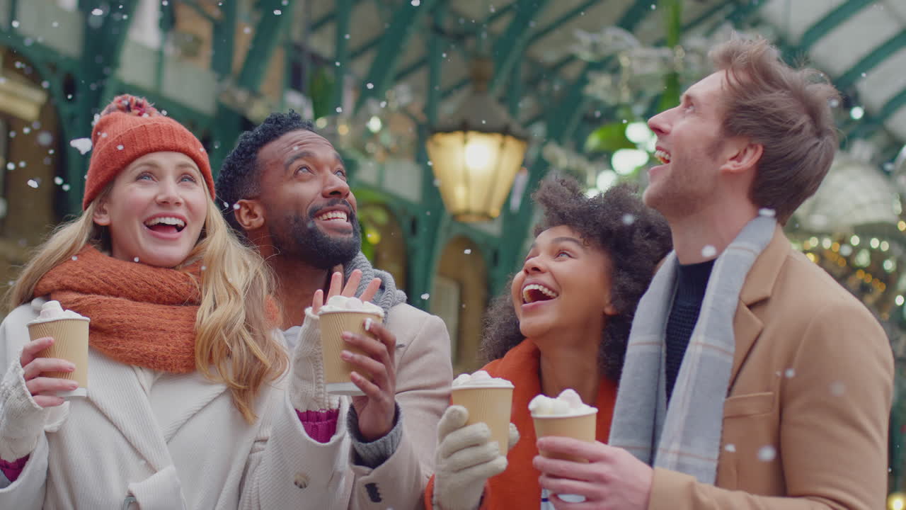 Group of friends drinking hot chocolate with marshmallows in snow at outdoor Christmas market in London's Covent Garden - shot in slow motion
