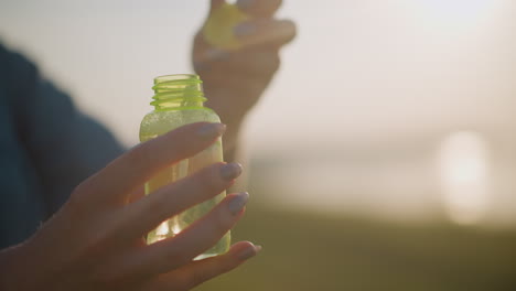 la mano de una mujer abre con gracia una pequeña botella espumosa bajo el suave resplandor de la luz solar, creando un ambiente cálido y pacífico en un entorno al aire libre. la luz mejora el momento sereno y delicado