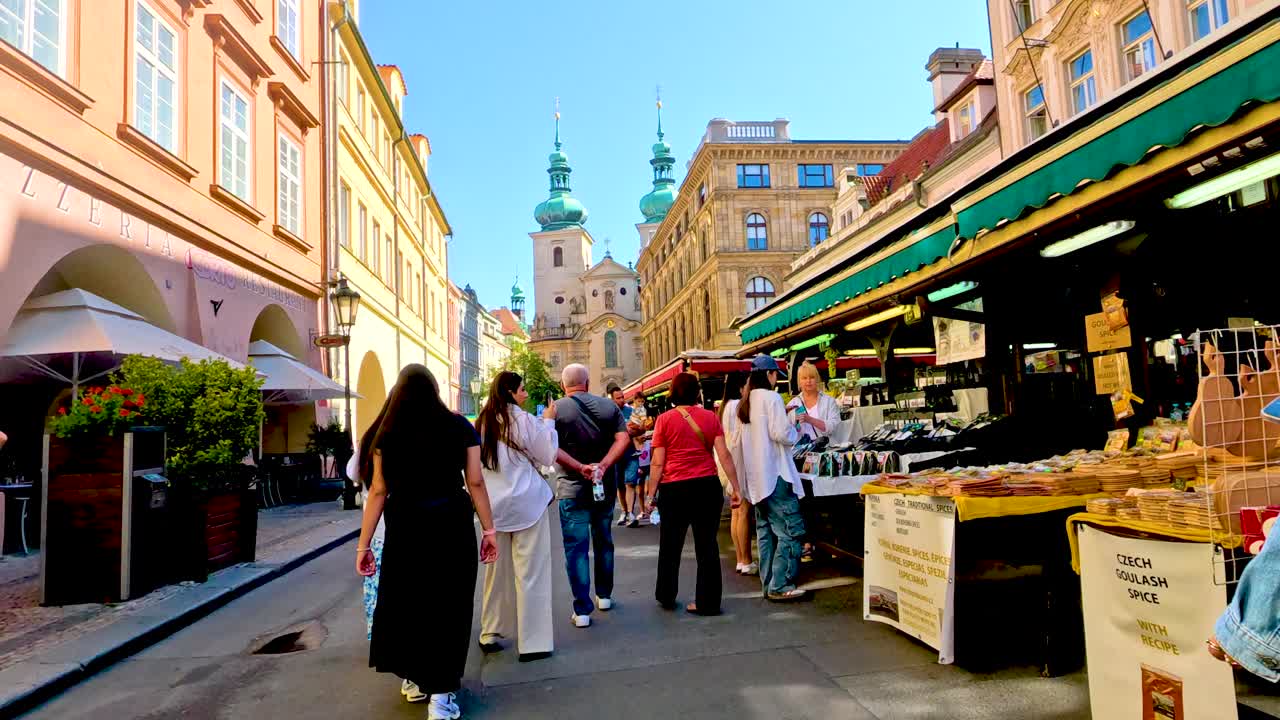 Lively pedestrians stroll past colorful market stalls in a sunlit historic Prague alleyway