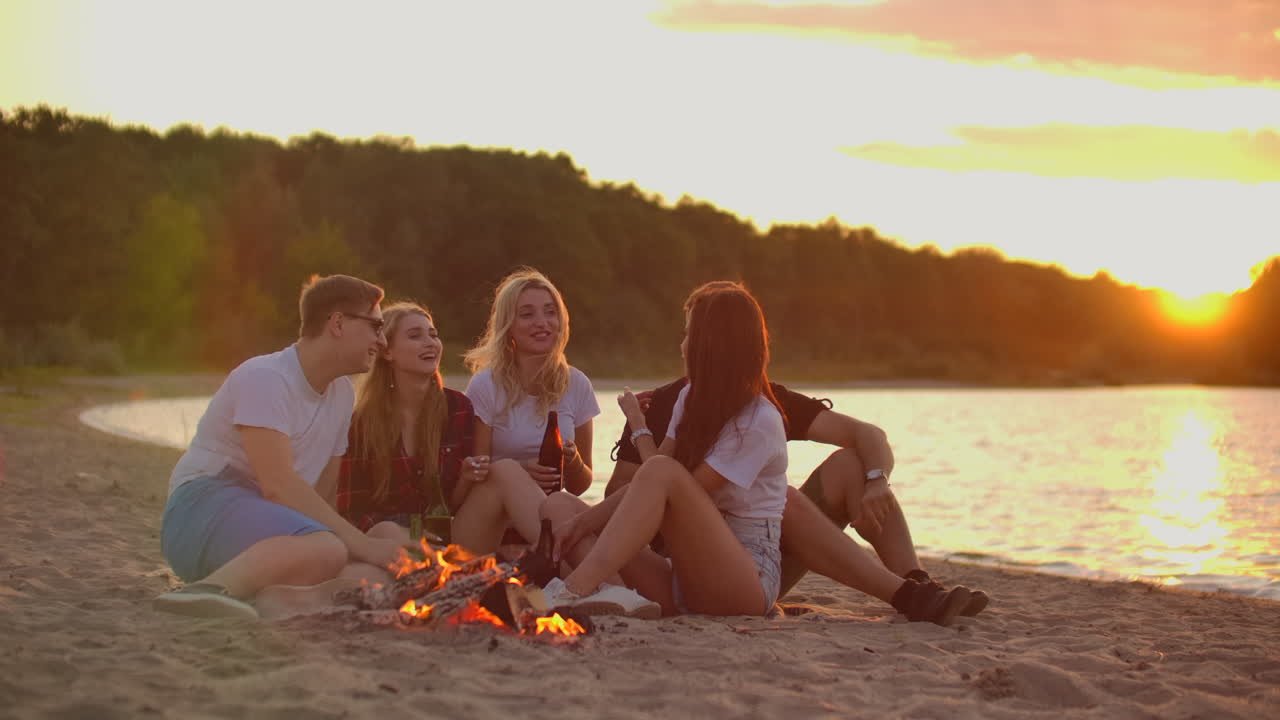The young blonde is telling a funny story to her friends around bonfire on the beach. They are drinking beer at sunset and enjoying the summer evening on the lake coast.