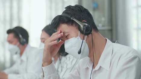 close up of a man of three asian call center agents wearing headset and mask headache to working while two of his colleagues are speaking and typing during the call with customer at the office