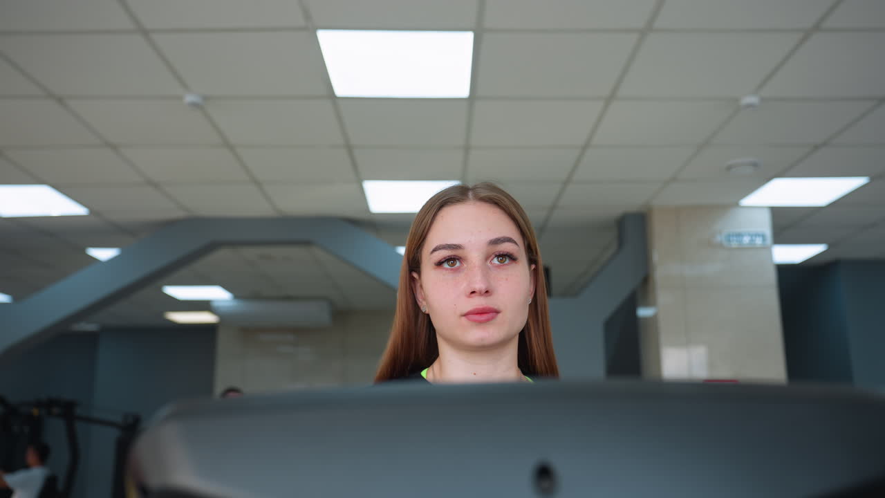 focused woman exercising on treadmill in modern indoor gym with bright ceiling lights and other people blurred in background, showcasing determination and healthy lifestyle in fitness environment