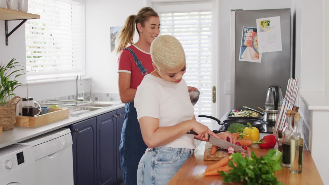 Casal feliz e diverso de mulheres cozinhando vegetais e se abraçando na cozinha.