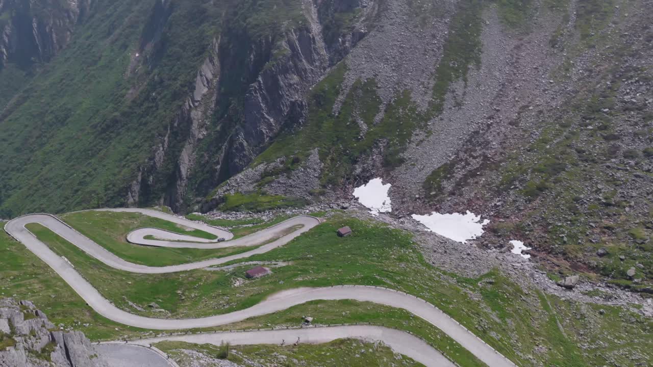 Alpine road switchbacks seen from above, curving through green and rocky cliffs