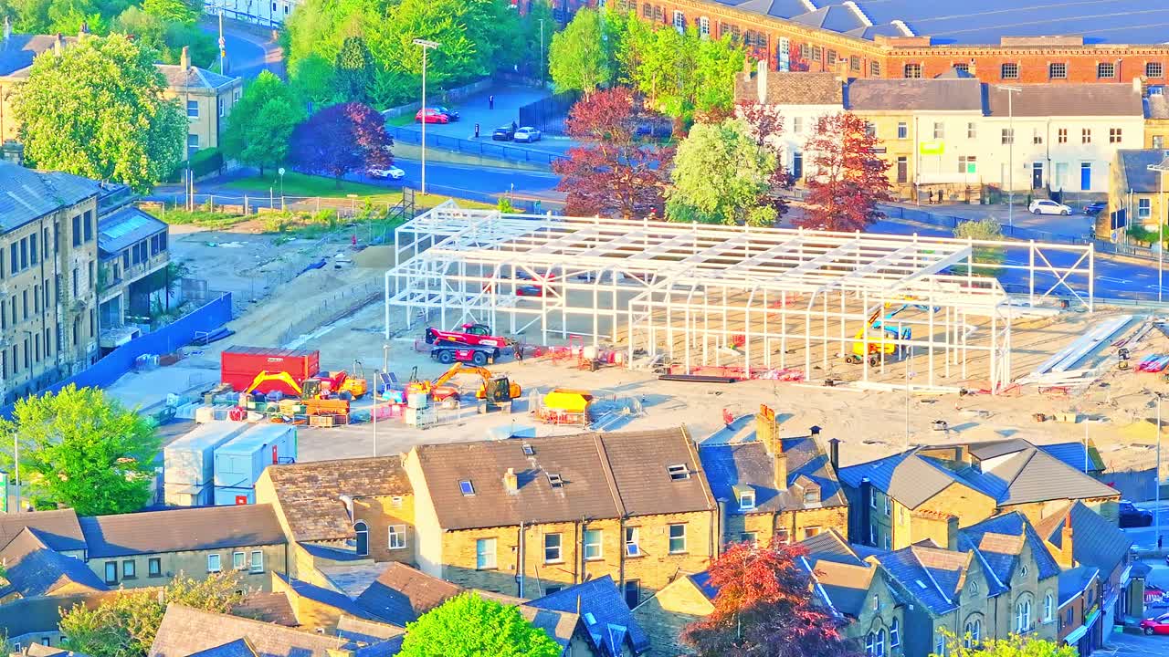 Huddersfield city centre revealing active construction site with steel framework being assembled, construction vehicles, nearby Victorian houses, trees, and local roads, drone static shot